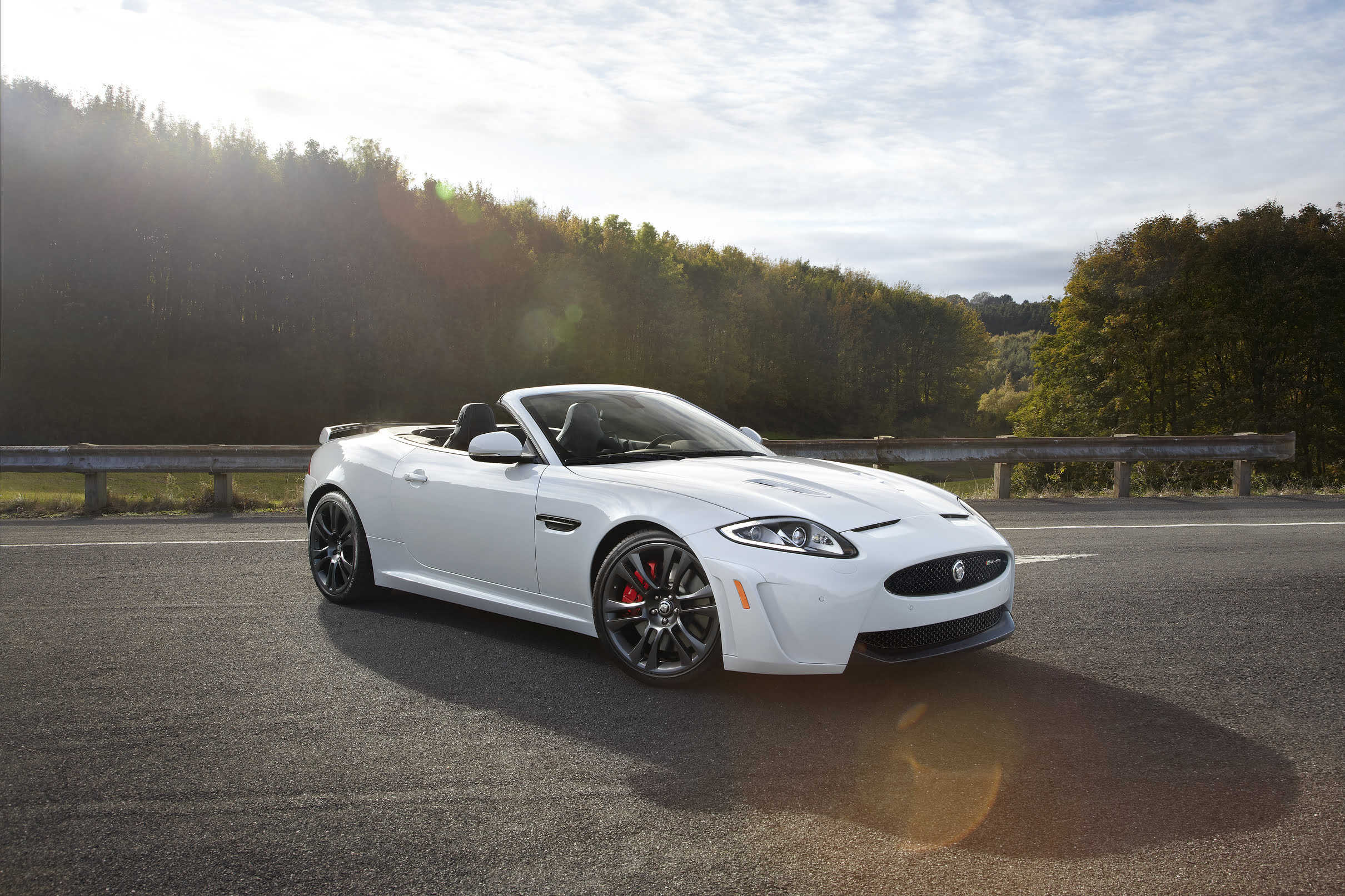 A black Jaguar XK convertible cruising along a scenic road, showcasing its sleek design and open-air driving experience in a luxurious setting.